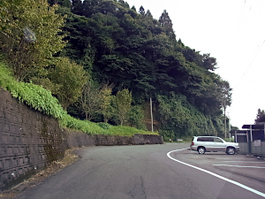 上り坂になっている中央線のない道、左手は山でコンクリートブロック擁壁が並び、右手は住宅の駐車場だ